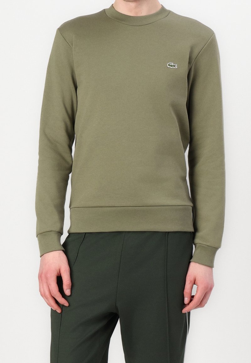 Homme portant un sweat-shirt à manches longues vert olive avec un petit logo crocodile sur la poitrine et un pantalon vert foncé, debout, les bras détendus.