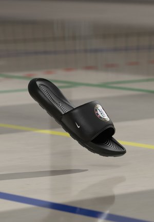 Sandale slide noire Nike avec le logo du Chelsea Football Club sur la sangle, flottant au-dessus d’un sol de terrain de sport avec des lignes de délimitation colorées.