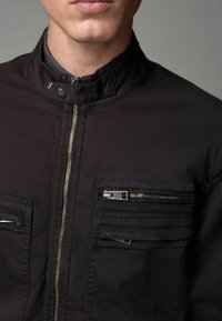 Zwarte jas met een hoge kraag, ritssluiting aan de voorkant en meerdere ritstasjes. De stof heeft een gladde textuur met verstevigde stiksels.
