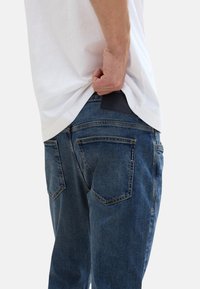 Sötétkék farmernadrág, slim fit szabással. Jellemzői közé tartozik a hátsó zsebek, a bőrpánt a derék részen és a kissé megkopott területek.