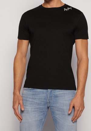 Mand iført enkel sort t-shirt med korte ærmer og hvidt "Replay"-logo på venstre skulder samt lyseblå denimjeans, stående foran en ensfarvet baggrund.