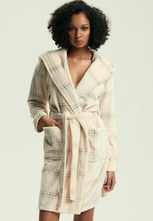 Woman wearing a soft, cream plaid robe with a wide collar, tied belt, and pockets, standing against a plain light background.