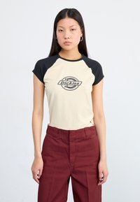 Eine junge Frau steht selbstbewusst da und trägt ein beige-schwarzes Kurzarmoberteil mit einem Logo, kombiniert mit weinroten Hochbundhosen.