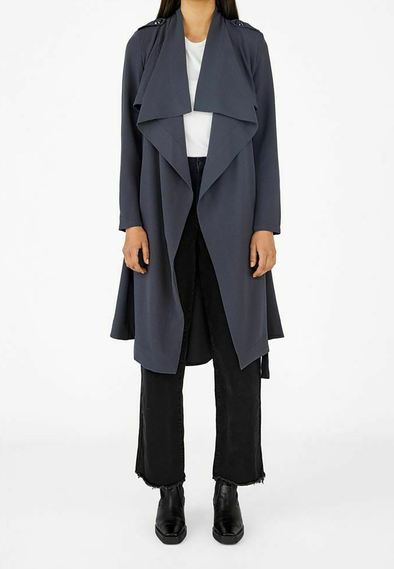 Femme portant un long trench-coat gris foncé ouvert sur le devant, chemise blanche, pantalon noir raccourci et bottines noires, debout sur fond blanc.