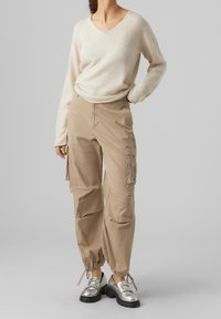 Beżowe spodnie cargo z elastycznymi mankietami i bocznymi kieszeniami, zestawione z kremowym swetrem w serek. Zestaw uzupełniają srebrne buty z odblaskowym wykończeniem.