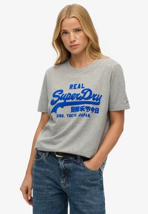 Szara bawełniana koszulka z krótkim rękawem z niebieskim nadrukiem logo "SuperDry". Okrągły dekolt, luźny krój, zestawiona z wysokim stanem ciemnych dżinsów.