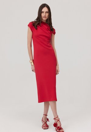 Kobieta w czerwonej sukience midi bez rękawów z marszczonym dekoltem i czerwonych kwiatowych sandałach na wysokim obcasie, stojąca na jednolitym tle.