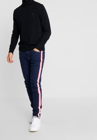 Maglione nero a collo alto abbinato a jeans blu scuro con strisce laterali bianche e rosse, completato da sneakers bianche.