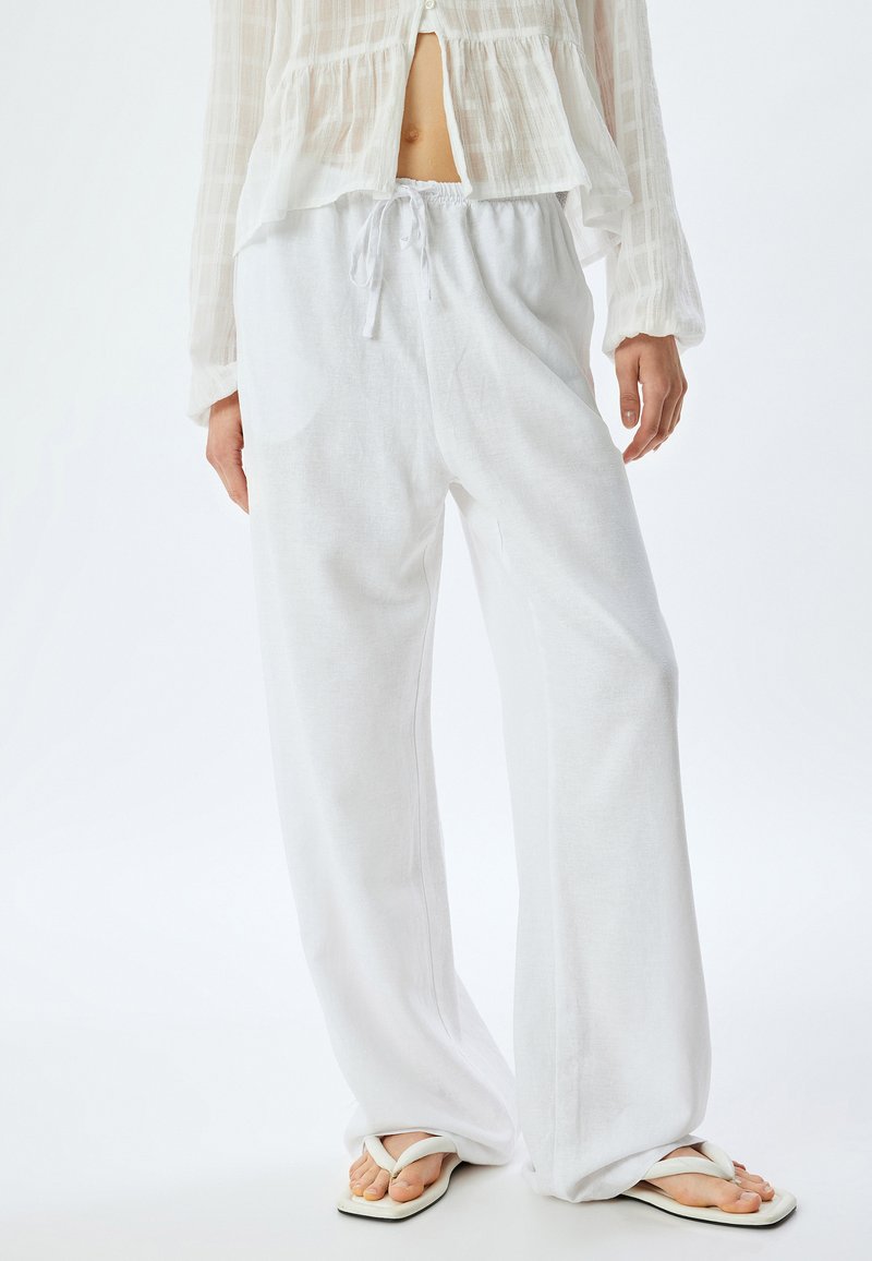 Witte linnen broek met een ontspannen pasvorm en elastische tailleband, voorzien van zijzakken en een lichte, luchtige textuur. Gecombineerd met witte slippers.