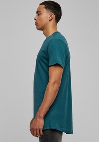 Tricou din bumbac de culoare teal, cu gât rotund, mâneci scurte și o croială lejeră. Materialul pare neted, cu o ușoară strălucire.