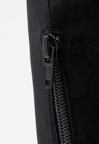 Prenda negra con cremallera que presenta un tejido suave y una textura delicada, con cremallera YKK. Incluye detalles de costura a lo largo de las costuras.