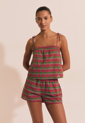 Kobieta stojąca, ubrana w czerwoną i zieloną kratę bez rękawów z wiązanymi ramiączkami na ramionach i dopasowane szorty na jednolitym beżowym tle.