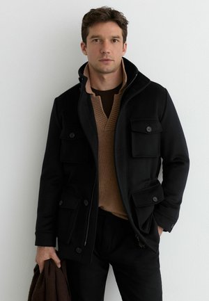 Sort yderjakke med flere forlommer foran, lynlås og høj krave, lagdelt over en brun tekstureret sweater.