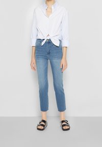 Camisa azul claro de botones anudada en la cintura, combinada con jeans ajustados de lavado medio y sandalias negras de tiras.