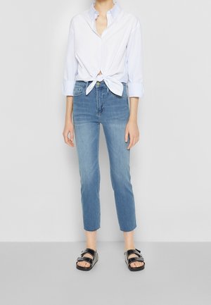Camicia azzurra chiara annodata in vita, abbinata a jeans attillati di media lavatura e sandali neri con cinturini.