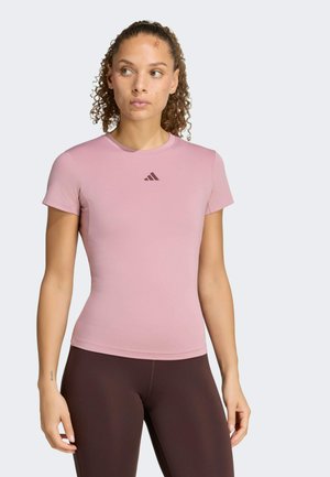 Donna con capelli ricci che indossa una maglietta aderente rosa chiaro a maniche corte con un piccolo logo scuro e leggings marroni scuri, in piedi davanti a uno sfondo uniforme.