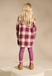 Ruudullinen flanellipaita vaaleanpunaisena ja kermana, ylisuuri istuvuus. Purppurat legginsit, kuvioidut sukat ja vaaleanruskeat kengät täydentävät asukokonaisuuden. Yksinkertainen tausta.