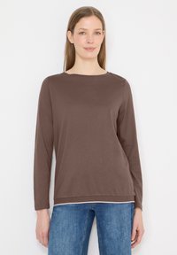 Femme aux longs cheveux châtain clair, portant un t-shirt marron uni à manches longues et un jean bleu, sur fond blanc.