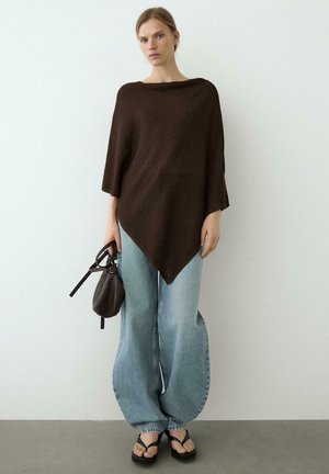Femme portant un poncho asymétrique marron foncé, un jean bleu clair à jambes larges, des sandales compensées noires, tenant un petit sac à main sombre.