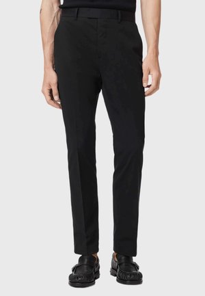 Mann trägt schwarze Slim-Fit-Anzughose mit schwarzem Gürtel und schwarzen Slippern, steht vor einem hellen einfarbigen Hintergrund.