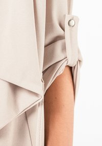 Lichtbeige blouse met een gedrapeerd ontwerp, zachte stoftextuur, opgerolde mouwen met knoopaccenten en een zijritsdetail.