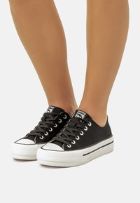 Zapatillas de lona negras con punta y suela de goma blanca, con cordones y ojales negros, lengua de marca y franja de contraste.