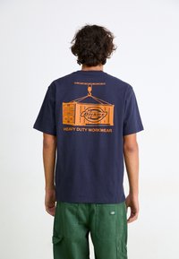 Temno mornarsko modra bombažna majica s kratkimi rokavi z oranžno grafiko, ki prikazuje žerjav, ki dviga kontejner, ter besedilom "Dickies HEAVY DUTY WORKWEAR."