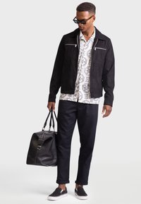 Black suede jacket with double front zippered pockets, white patterned shirt, black trousers, and black sneakers, holding a black leather bag.