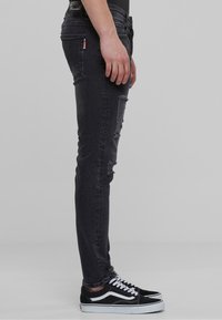 Jeans de mezclilla negros ajustados con parches desgastados, forma de pierna estrecha y una sutil textura deslavada, combinados con zapatillas negras.