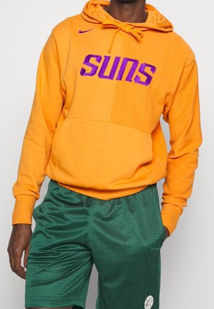 Personne portant un sweat à capuche orange avec le texte violet "SUNS" et un short en maille vert avec un logo blanc, debout avec une main dans la poche.