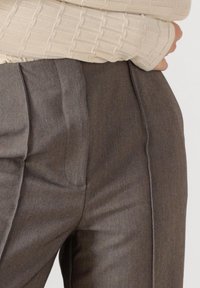 Zbliżenie osoby w szarych, dopasowanych spodniach i beżowej, teksturowanej bluzce z długim rękawem, z ręką spoczywającą na talii.