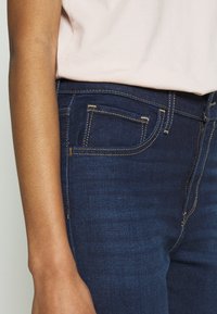 Tmavě modré džíny s žlutým prošíváním, malou přední kapsou a přiléhavým střihem, nošené s světle růžovým topem.