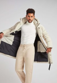Giacca puffer beige con cappuccio, chiusura con zip e tasche interne, abbinata a pantaloni da jogging beige chiaro. Texture liscia, design minimalista.