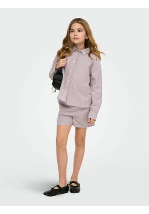 Mlada deklica stoji, oblečena v ujemajočo se dolgo majico na črte in kratke hlače, drži črno torbico čez ramo ter nosi črne čevlje Mary Jane.