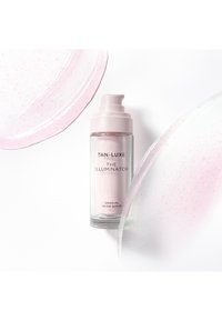 Flacon en verre transparent avec pompe rose contenant le sérum au glow progressif Tan-Luxe The Illuminator, entouré de traces de sérum teintées de rose.