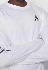 Person, der ein weißes Langarmshirt mit verschränkten Armen trägt, schwarzes geometrisches Logo auf der Brust und schwarzes Krallenmuster am rechten Ärmel.