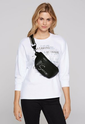 Schwarze Umhängetasche mit einem Reißverschlussfach und weißem Logo. Glatte Ledertextur, schräge Form, getragen über einem weißen Sweatshirt und schwarzen Jeans.