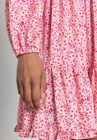 Vaaleanpunainen kukkamekko, jossa on röyhelöhelma, ja se on valmistettu tekstuuripinnasta, joka on peitetty pienillä, eloisilla kukilla vaaleanpunaisena, keltaisena ja valkoisena.