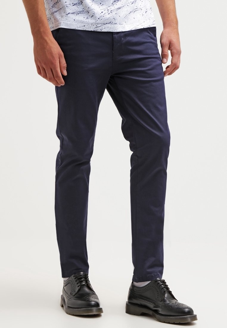 Muž v námořnických slim-fit kalhotách, černých kožených brogue botách a bílém vzorovaném tričku, stojící na pozadí jednobarevného světlého pozadí.