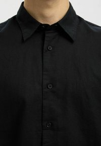 Gros plan d'une personne portant une chemise noire à boutons avec un col, montrant trois boutons visibles et la partie supérieure de la poitrine.