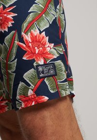 Pantaloni da bagno floreali blu con fiori rossi e bianchi, foglie verdi e un'etichetta nera "Superdry" cucita sul lato.