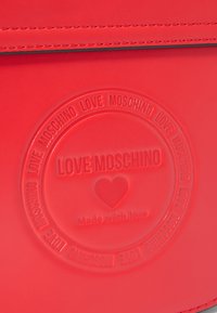 Červená taška z umělé kůže s vyvýšeným kruhovým logem a textem "LOVE MOSCHINO" a ikonou srdce, doplněná kontrastním prošíváním.