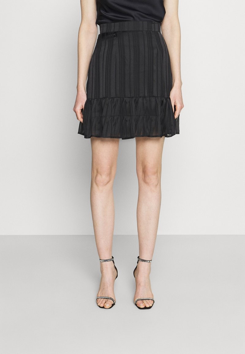 Jupe plissée noire avec un ourlet à volants, en tissu léger, présentant des rayures verticales et une ceinture ajustée. Sandales noires à brides.