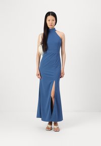 Rochie maxi cu fante în albastru, având un guler înalt și fante pe lateral. Modelul ține o geantă mică albă și poartă sandale cu barete bej. Textură netedă.