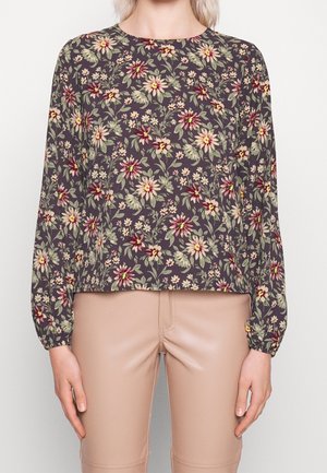 Vrouw draagt een lange mouw donker bloemig topje met beige high-waist broek, handen ontspannen langs het lichaam tegen een effen achtergrond.