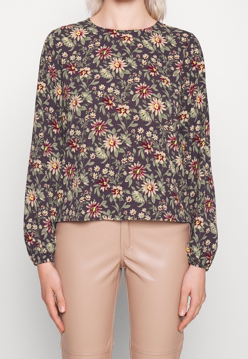 Femme portant un haut à manches longues à motif floral sombre avec un pantalon beige taille haute, les mains détendues sur les côtés devant un fond uni.