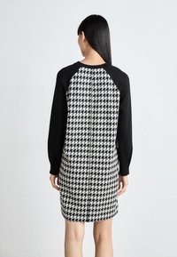 Mustavalkoinen houndstooth-mekko pitkilla hihoilla, kontrastina mustat hihat ja pyöreä pääntie. Sileä tekstuuri, polvipituinen.