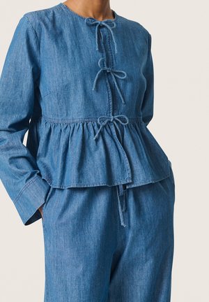 Kvinde iført en blå langærmet denimtop med tre bundne sløjfer ned foran og matchende løstsiddende denimbukser.