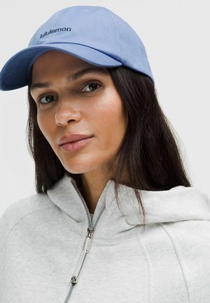 Kobieta w jasnoniebieskiej czapce Lululemon i jasnoszarej bluzie z suwakiem, spoglądająca lekko na bok z neutralnym wyrazem twarzy.