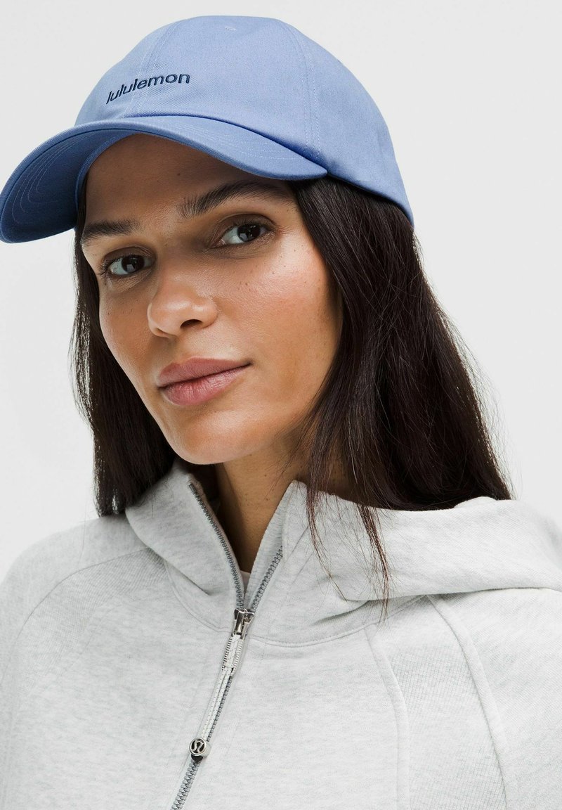 Frau mit einer hellblauen Lululemon-Kappe und einem hellgrauen, mit Reißverschluss versehenen Hoodie, die leicht seitlich blickt mit einem neutralen Gesichtsausdruck.